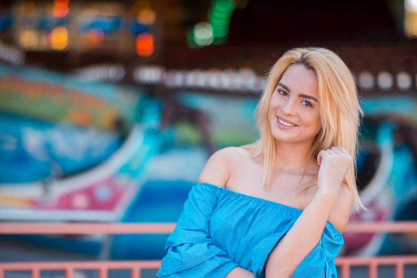 Happy young woman with blond hair in bright blue top smiling against the backdrop of a carousel. Summer portrait of a girl on a bright background - Photo, Image