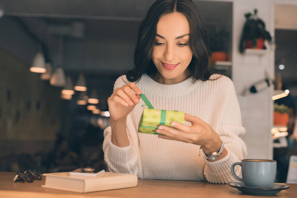 smiling young woman untying gift box at table with smartphone and coffee cup in cafe - Photo, Image