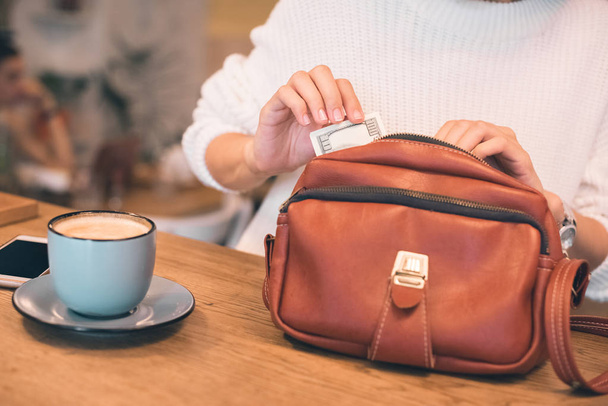 cropped image of woman pulling out cash from bag at table with coffee cup in cafe  - Photo, Image