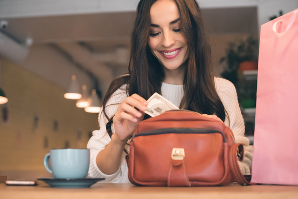 smiling woman pulling out cash from bag at table with coffee cup in cafe  - Photo, Image
