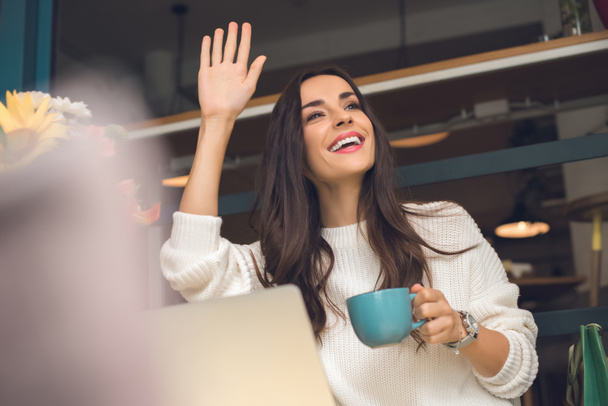 feliz freelancer femenina con taza de café saludando a mano en la mesa con el ordenador portátil en la cafetería
 - Foto, imagen