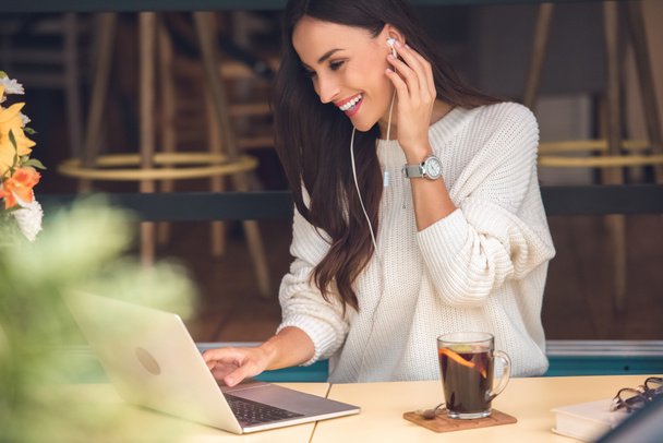 szczęśliwy młodych kobiet freelancer w słuchawki pracy na laptopie przy stole z grzanym winem w kawiarni  - Zdjęcie, obraz