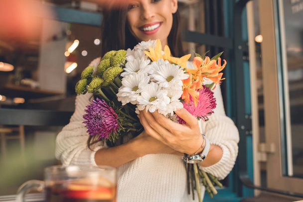 selectieve aandacht van gelukkige jonge vrouw houdt van kleurrijke boeket uit diverse bloemen in café - Foto, afbeelding