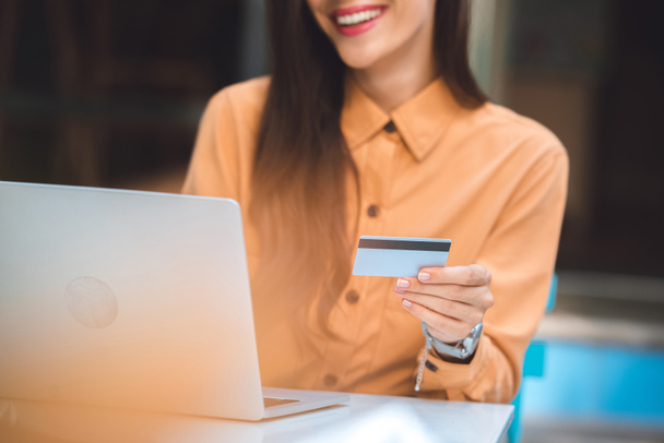 частковий вид на стильну жінку з кредитною карткою, що робить онлайн покупки на ноутбуці за столом у кафе на міській вулиці
 - Фото, зображення