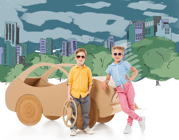 stylish children in sunglasses posing near cardboard car in drawn city and park - Photo, Image