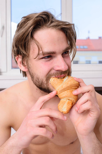 Enjoy every bite. Man eats croissant he likes bakery products. Guy bites sweet dessert. Man bearded handsome guy eating breakfast in bed. Guy holds croissant bedroom hotel room window background - Фото, изображение