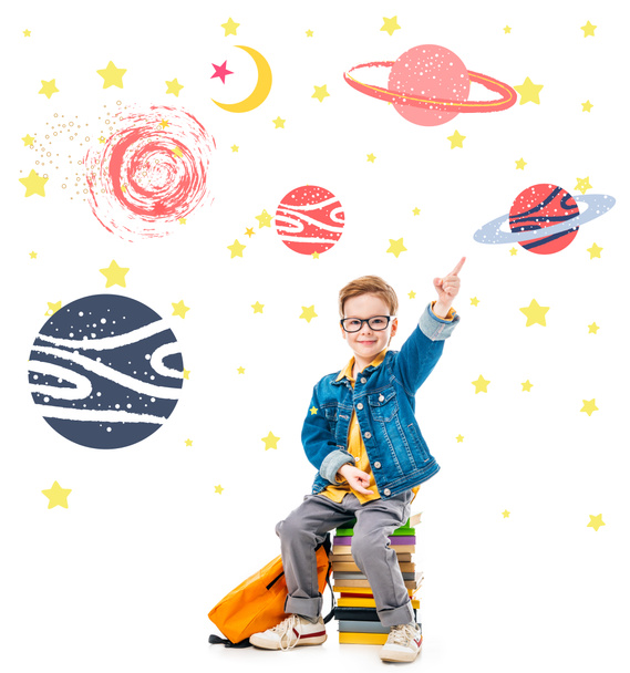 smiling schoolboy pointing up while sitting on pile of books with backpack, isolated on white with fantasy universe and planets - Foto, afbeelding