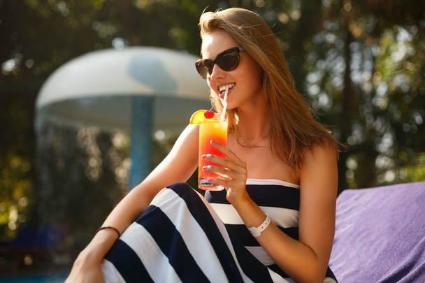 Portrait of young woman with cocktail glass near swimming pool on a deck chair with palm trees behind. Vacation concept - Foto, Imagem