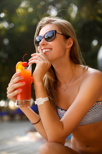 Portrait of young woman with cocktail glass chilling in the tropical sun near swimming pool on a deck chair with palm trees behind. Vacation concept - Photo, Image