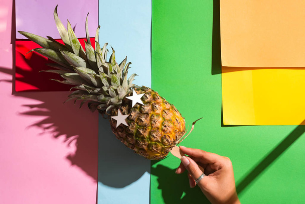 Naispuolinen käsi pitää jäätelöä kiinni ananaksella jäätelönä värillisellä taustalla. Ananassilmät valkoisten puisten tähtien muodossa. Juhlaananas
 - Valokuva, kuva