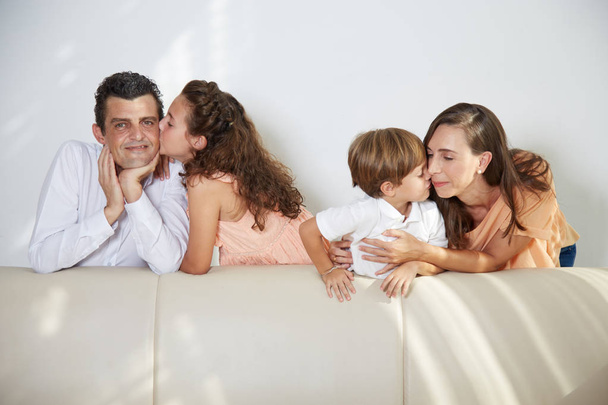Ευτυχισμένα παιδιά φιλιά γονείς στα μάγουλα, στο σπίτι κοντά στο καναπέ  - Φωτογραφία, εικόνα