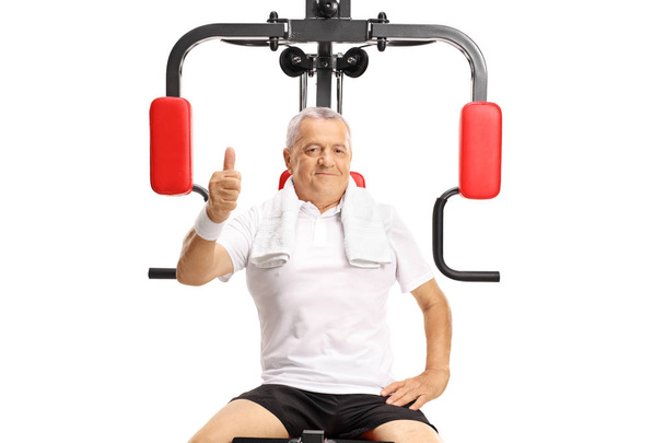 Mature man seated on an exercise machine holding his thumb up isolated on white background - Foto, Imagem
