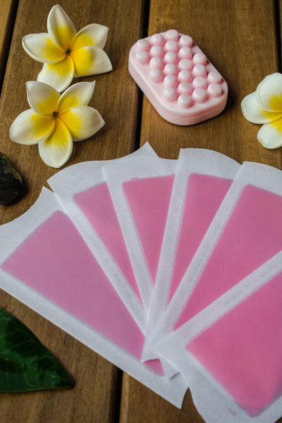 Pink cold wax strips with pink soap bar on wooden surface with plumeria flowers and black round stones - Photo, Image