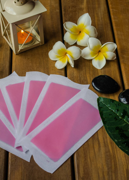Pink cold wax strips  on wooden surface with plumeria flowers, orange candle  and black round stones - Photo, Image