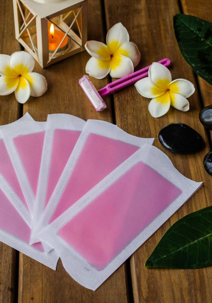 Pink cold wax strips with pink razor blade on wooden surface with plumeria flowers and black round stones - Photo, Image