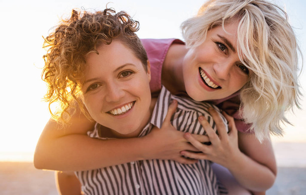 Portrait of a smiling young woman carrying her laughing girlfriend on her shoulders while enjoying an afternoon together at a beach  - Photo, image