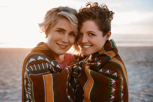 Portrait of a smiling young lesbian couple wrapped in a warm blanket while enjoying the sunset together at the beach - Photo, Image