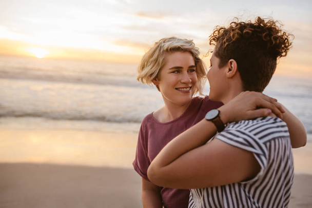 Улыбающаяся молодая лесбийская пара, стоящая рука об руку на песчаном пляже и любующаяся закатом
  - Фото, изображение