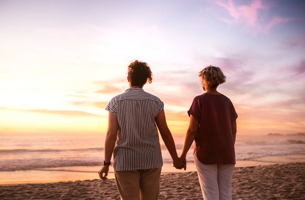 Rückansicht eines jungen lesbischen Paares, das Hand in Hand an einem Sandstrand steht und einen romantischen Sonnenuntergang über dem Ozean beobachtet - Foto, Bild