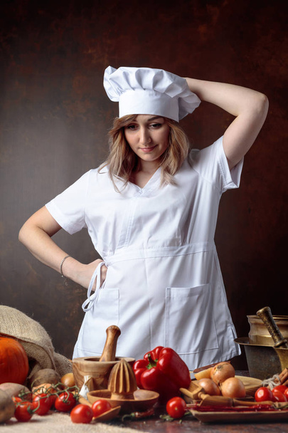 Fiatal gyönyörű lány egy egységes szakács azt hiszi, előtt egy tábla különböző konyhai eszközök és zöldségekkel. - Fotó, kép