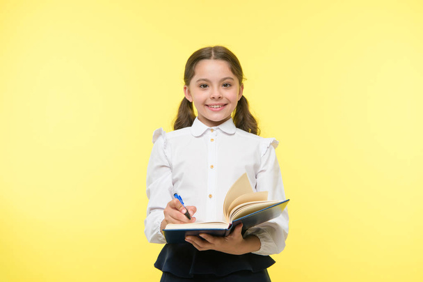 Write note to remember. Child school uniform smart kid happy make note. Girl happy face make note about idea yellow background. Child girl happy school uniform clothes holds book ready write note - Φωτογραφία, εικόνα