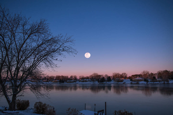 Cerca de Luna Llena Levantándose sobre el Lago y Casas en Cielo Rojo y Azul de Invierno
 - Foto, imagen