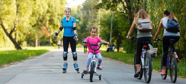 молодая мама катается на роликах с маленькой дочерью на велосипеде в парке
 - Фото, изображение