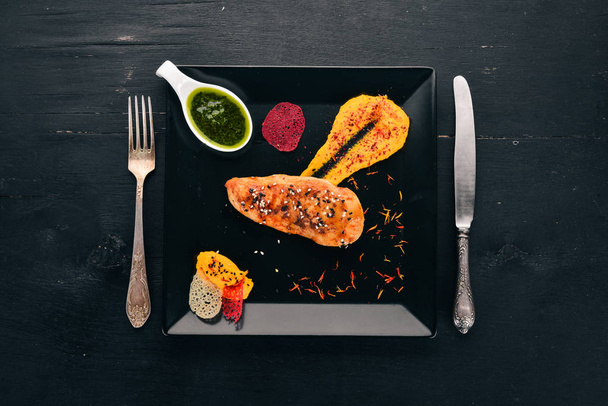 Baked chicken fillet with sauce on a black plate. On a wooden background. Free space for text. Top view. - Foto, afbeelding
