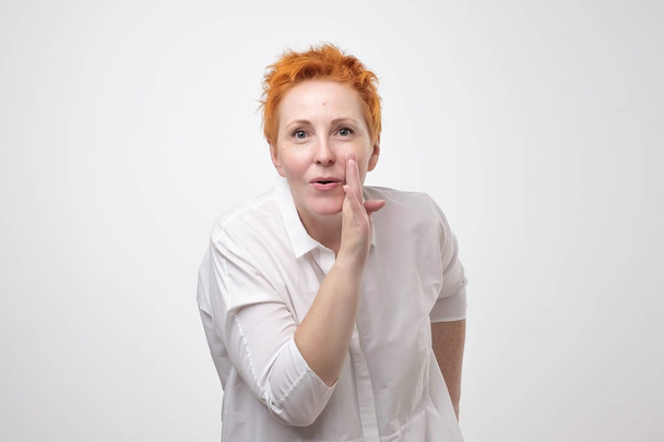 Do not tell anyone concept. Talkative prettyeuropean woman with red hair is saying secret hot braking news isolated on grey background - Photo, Image
