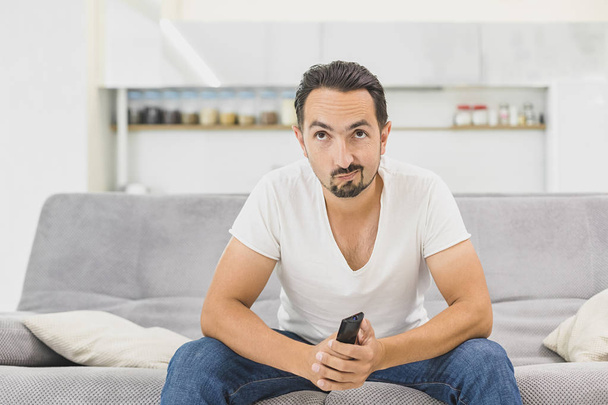 Nothing interesting to watch. Handsome young man holding remote control and looking bored while watching TV on the couch at home - Photo, Image