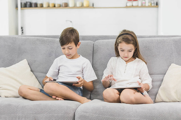 Two kids with gadgets. Sister and brother surfing the net or playing online games on digital tablets at home. Modern communication and gadget addiction concept - Zdjęcie, obraz