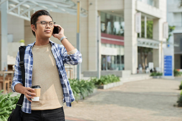 Χαμογελώντας Φιλιππίνων νεαρός άνδρας μιλά στο τηλέφωνο και κρατώντας καφέ, ενώ το περπάτημα στο δρόμο - Φωτογραφία, εικόνα