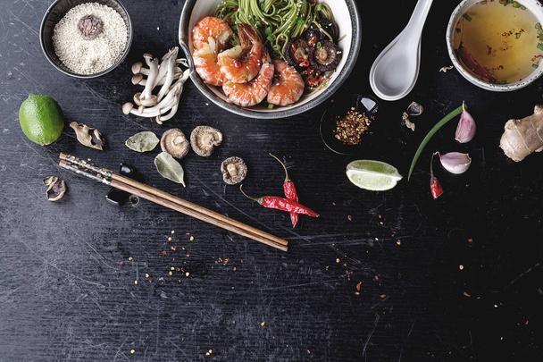Green tea soba noodles  served with shrimps,  shiitake mushrooms and spicy broth.  Decorated with different spices and condiments used during preparation: garlic, lime, chili pepper, shimeji mushrooms, ginger, lime leaves, and sesame seeds. - Photo, Image