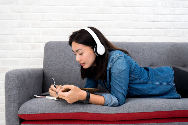 Woman listening to music and writing diary on sofa at home - Foto, afbeelding