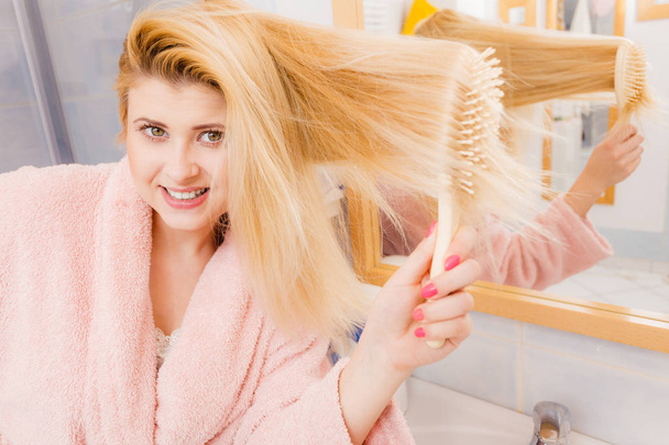 Woman wearing dressing gown brushing her long blonde hair, morning beauty routine. Haircare and hairstyling concept. - Photo, image