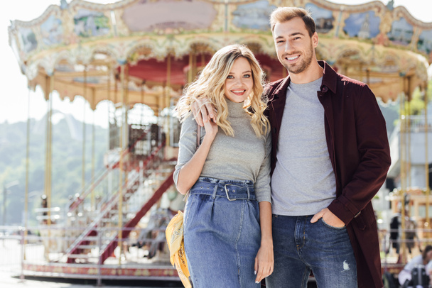 smiling affectionate couple in autumn outfit cuddling near carousel in amusement park and looking at camera - Foto, Bild