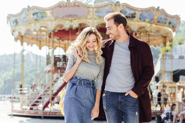 smiling couple in autumn outfit walking and hugging near carousel in amusement park - Фото, изображение