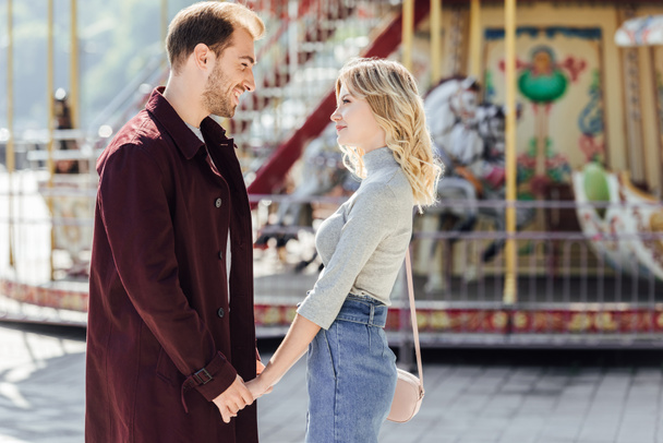 aanhankelijk paar in herfst-outfit hand in hand en kijken elkaar in de buurt van carrousel in attractiepark - Foto, afbeelding