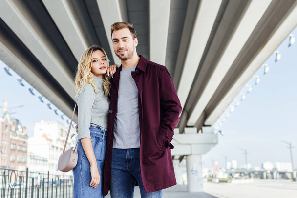 stylish couple in autumn outfit hugging under bridge in city and looking at camera - Foto, Imagem