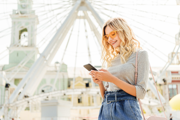 красивая улыбающаяся женщина в осеннем наряде с помощью смартфона возле колеса наблюдения в городе
 - Фото, изображение
