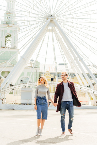 romantic couple in autumn outfit holding hands and walking near observation wheel - Фото, изображение