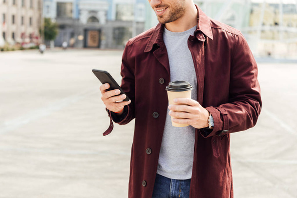 cropped image of man in autumn outfit holding coffee to go and using smartphone in city - 写真・画像