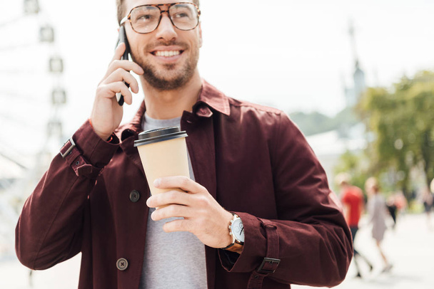 smiling man in autumn outfit holding coffee to go and talking by smartphone in city - Photo, Image