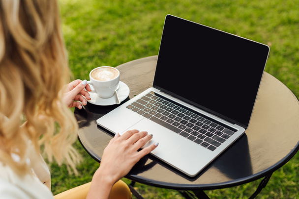 abgeschnittenes Bild einer Frau, die Laptop mit leerem Bildschirm benutzt und eine Tasse Kaffee auf einem Tisch im Garten hält - Foto, Bild