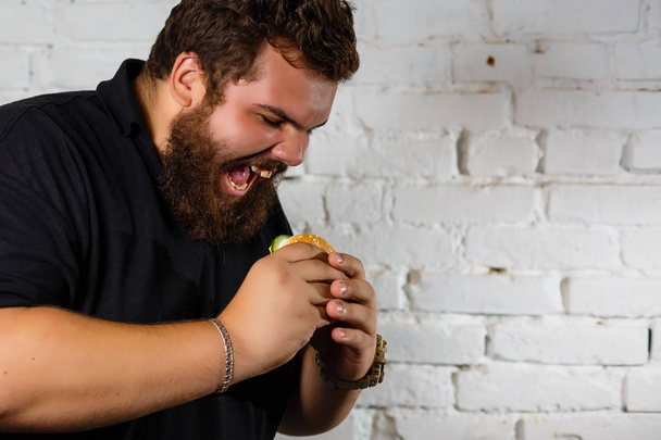 Fat Man λαίμαργα τρώει χάμπουργκερ σε φόντο λευκό τούβλο τοίχο - Φωτογραφία, εικόνα