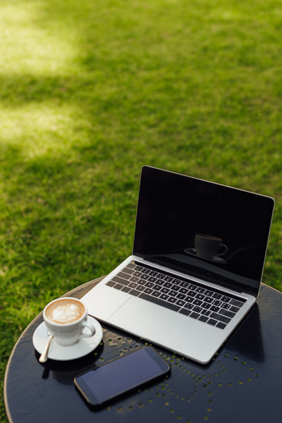 Laptop und Smartphone mit leeren Bildschirmen und einer Tasse Cappuccino auf dem Tisch im Garten  - Foto, Bild