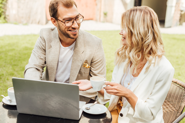 χαμογελώντας ζευγάρι στο φθινόπωρο στολή κάθεται στο τραπέζι με το laptop και πίνοντας τον καφέ στην καφετέρια - Φωτογραφία, εικόνα