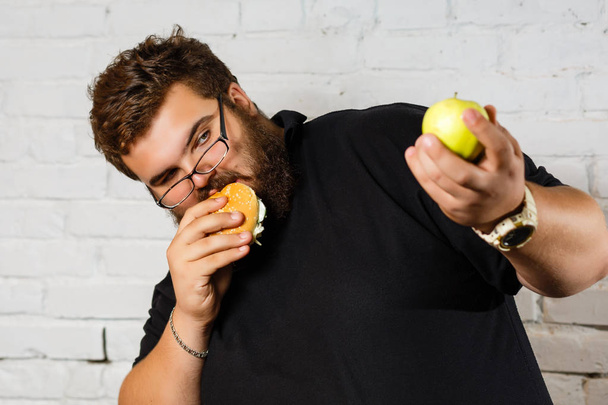 Excited bearded man greedily eating hamburgers and holding green apple in another hand on white background - Φωτογραφία, εικόνα