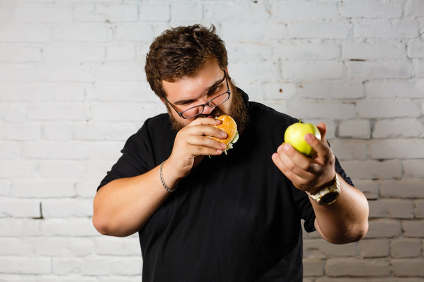 aufgeregter bärtiger Mann isst gierig Hamburger und hält grünen Apfel in einer anderen Hand auf weißem Hintergrund - Foto, Bild