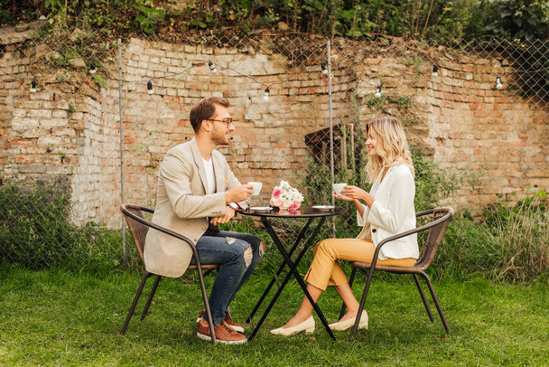 vista lateral de la pareja sentada en la mesa en la cafetería con tazas de café y ramo de flores en la mesa
 - Foto, Imagen
